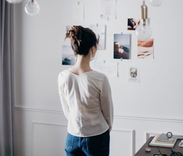 A woman standing with her back to the camera, looking at the wall in front of her which has photos taped to it. The room is white with a desk in the corner.